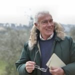 Happy senior man in outdoor setting holding glasses and a book, smiling warmly.